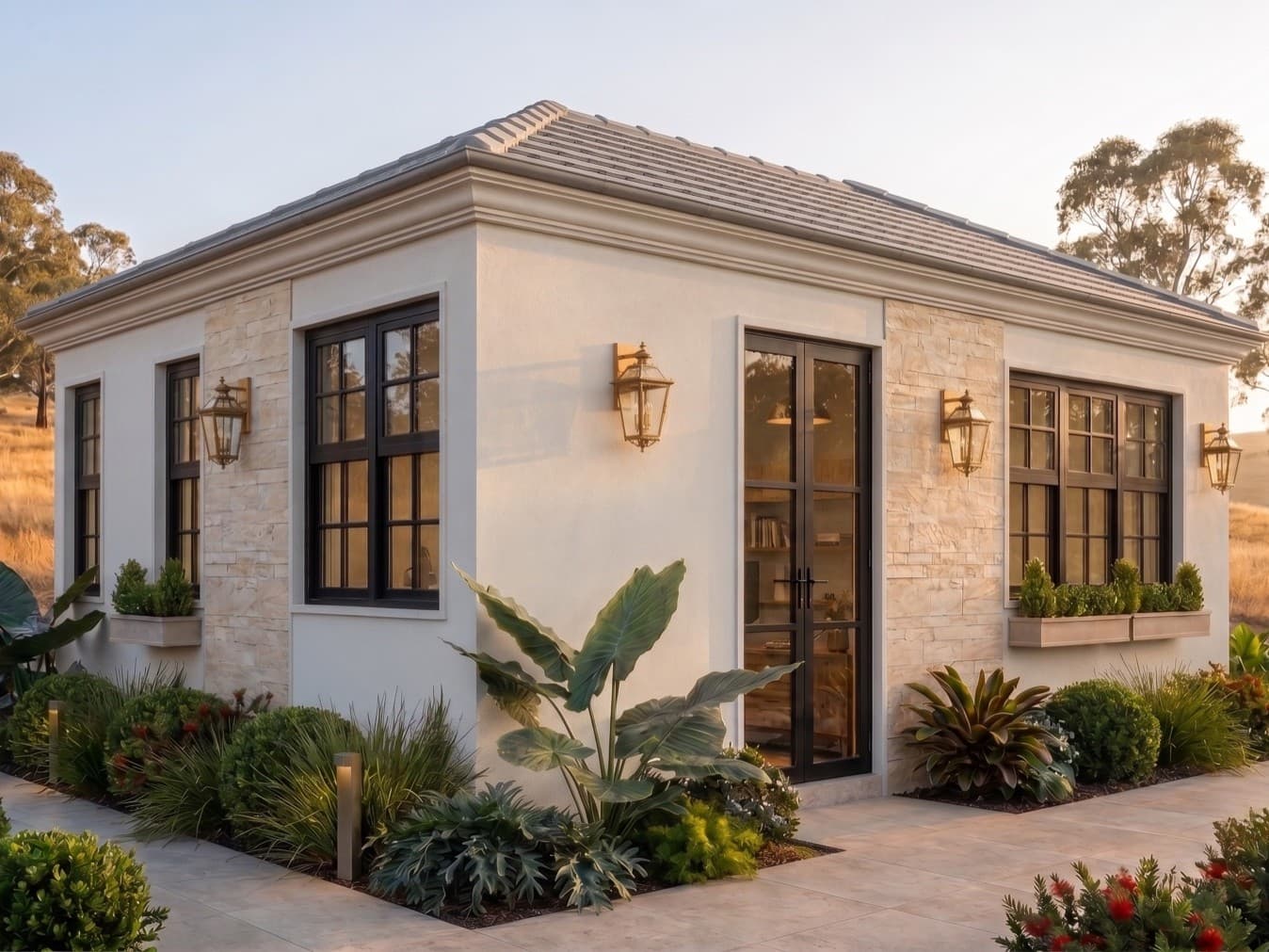 Artist's impression — the Elementa one-bedroom home in late-afternoon sun: off-white rendered walls with stone-clad panels, black-framed windows and glass doors, brass lanterns and planter boxes under a hip roof, in a planted garden with gum trees and golden grass behind.